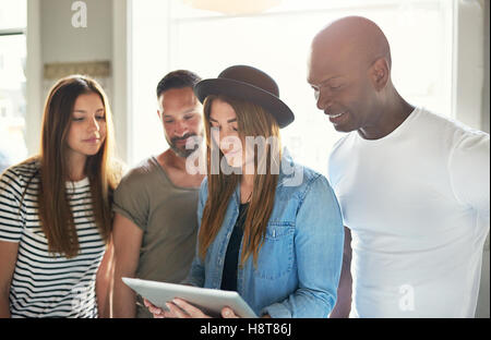 Gruppe von vier jungen Erwachsenen gekleidet als Studenten Tablet mit hellen Büro oder Schule Fenster dahinter zu betrachten Stockfoto