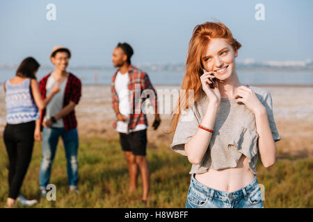 Fröhliche Rothaarige junge Frau am Handy-Stand in der Nähe ihrer Freunde im freien Stockfoto