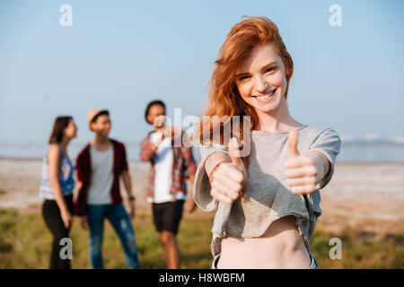 Fröhliche Rothaarige junge Frau stehen und zeigt Daumen hoch, während ihre Freunde im freien sprechen Stockfoto