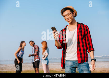Gerne asiatische junge Mann stehend und mit Handy während seiner Freunde im freien sprechen Stockfoto