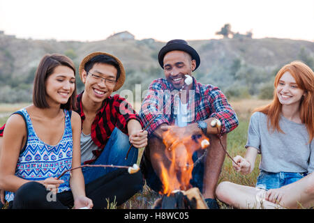 Gruppe der glückliche junge Menschen sitzen und Rösten Marshmallows am Lagerfeuer im freien Stockfoto