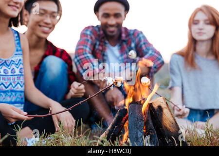 Multiethnische Gruppe von fröhlichen Jugendlichen sprechen und Marshmallows am Lagerfeuer im Freien im Sommer vorbereiten Stockfoto