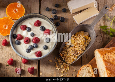 Ein gesundes Frühstück auf einem rustikalen Tisch mit Müsli, Joghurt, Himbeeren, Heidelbeeren, Mandarinen, Brot und Käse. Stockfoto