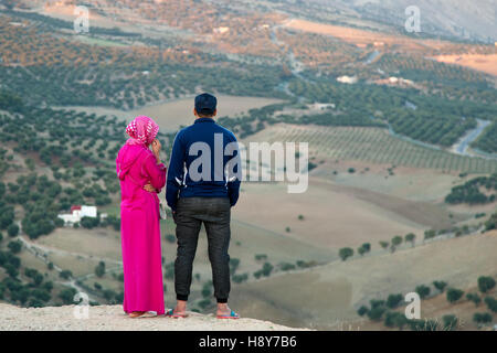 Ein Paar blickt von einem Hügel in der Nähe von Fès, Marokko, über ein Tal mit weitläufigen Olivenhainen, die Frau trägt einen rosa Kaftan, der Mann eine blaue Jacke. Stockfoto