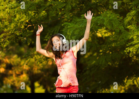 Glücklich Teenager-Mädchen in rot tanzen in der Natur beim Musikhören über Kopfhörer, lächelnd und Blick in die Kamera, an einem sonnigen Sommertag. Stockfoto