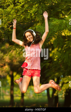 Glücklich Teenager-Mädchen im roten Sprünge im Park beim Musikhören über Kopfhörer, lächelnd und Blick in die Kamera, an einem sonnigen Sommertag. Stockfoto