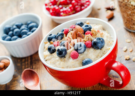 Haferflocken-Porridge mit frischen Heidelbeeren, Himbeeren, Müsli und Mandeln in der roten Schale am weißen Tisch. Gesundes Frühstück Stockfoto