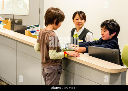 Japan, Nishinomiya, KidZania, Kind activity center. Bank, Interieur, Kind, Junge, Abhebungen vom Cashier mit jungen erwachsenen Frau überwachen. Stockfoto