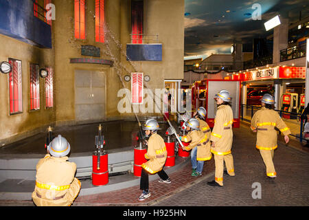 Japan, Nishinomiya, KidZania, untergeordnete Aktivität spielen. Leitung von Kindern als Feuerwehrleute, die Schläuche am brennenden Gebäude gekleidet. Stockfoto