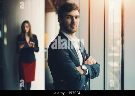 Gruppe von Geschäftsleuten in Büroeinrichtung: ernster Mann in formellen Anzug vor und Frau im roten Kleid und Jacke hinter, Mann stand in der Nähe von Fenster Stockfoto