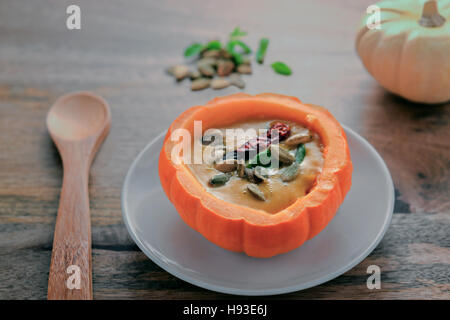 Kürbis-Suppe serviert in Schale aus Mini-Kürbis auf Holztisch, garniert mit Majoran, Kürbiskerne und rote Paprika, bereit mit Woode gegessen werden Stockfoto