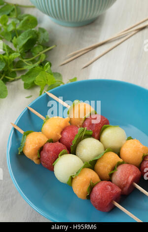 Obstspieße, Wassermelone, Melone, Galia Melone und Minze Blätter gemacht. Aufgereiht auf einem blauen Teller und frische Minzblätter im Hintergrund. Stockfoto