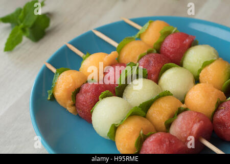 Obstspieße, Wassermelone, Melone, Galia Melone und Minze Blätter gemacht. Aufgereiht auf einem blauen Teller und frische Minzblätter im Hintergrund. Stockfoto