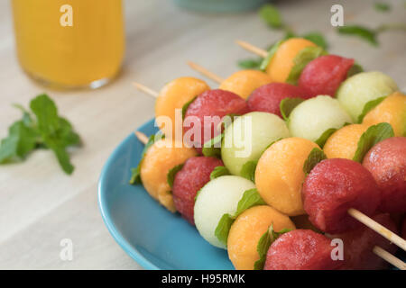 Obstspieße, Wassermelone, Melone, Galia Melone und Minze Blätter gemacht. Aufgereiht auf einem blauen Teller und frische Minzblätter im Hintergrund. Stockfoto