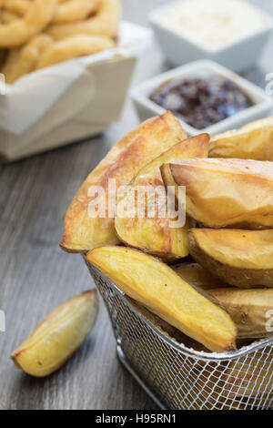 Fettfrei gebacken Kartoffelecken in einem Mini-Drahtkorb auf einem Holztisch mit Zwiebelringen, Barbeque-Sauce und Mayonnaise im Hintergrund. Stockfoto