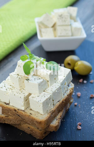 Tofu-Feta-Käse mit frischem Oregano auf Vollkorn Toast mit Leinsamen zusammen mit Himalaya-Salz und zwei grüne Oliven auf einem schwarzen Stein Tablett. Stockfoto