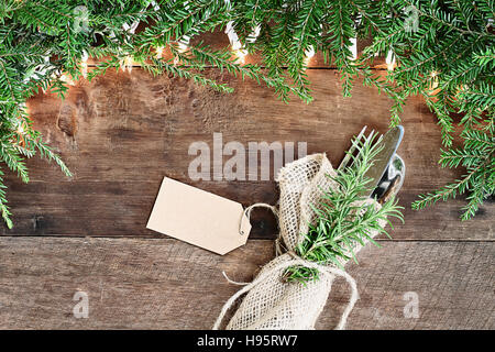 Weihnachtsbaum Tannenzweigen, Besteck in eine Serviette Sackleinen mit Grußkarte und dekorative Leuchten über einen rustikalen Hintergrund der Scheune Holz gewickelt. Stockfoto