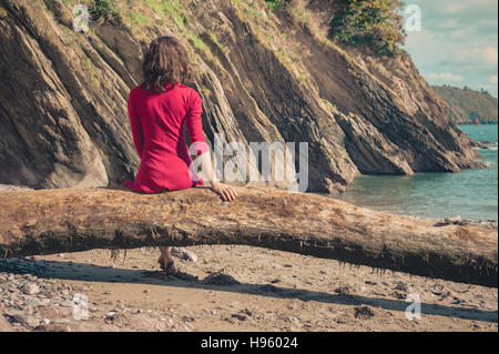 Eine junge Frau trägt ein rotes Kleid sitzt auf einem Baumstamm am Strand und ist entspannend Stockfoto