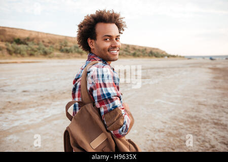Lächelnd afroamerikanischen jungen Mann mit Rucksack zu Fuß am Strand Stockfoto