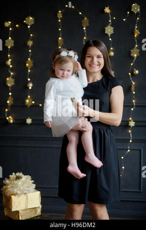 Junge Frau mit der kleinen Tochter auf Händen. Ruddy Baby trägt ein weißes Kleid, auf den Kopf von ihr einen Kranz nieder. Dunkle Wand schmückt sich mit gold g Stockfoto