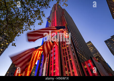 Rockefeller Center in der Dämmerung in weiß, rot und blau beleuchtet. Amerikanische Flaggen flattern im Wind. Midtown Manhattan, New York Stockfoto