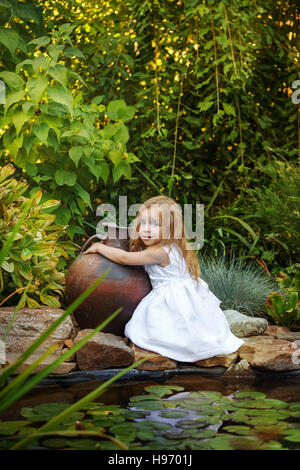 Niedliche kleine Mädchen in einem weißen Kleid hält eine große alte Tonkrug in den Seerosenteich. Stockfoto