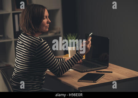 Business-Frau Überstunden im Büro, kaukasische Erwachsenfrau SMS von ihrem Smartphone beim Abschluss von Projektaufgabe auf Laptop com Stockfoto