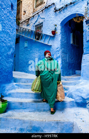 Eine ältere Frau Spaziergänge durch die bunten Gassen der Medina mit schweren Einkaufstüten, Chefchaouen, Marokko Stockfoto