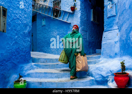 Eine ältere Frau Spaziergänge durch die bunten Gassen der Medina mit schweren Einkaufstüten, Chefchaouen, Marokko Stockfoto