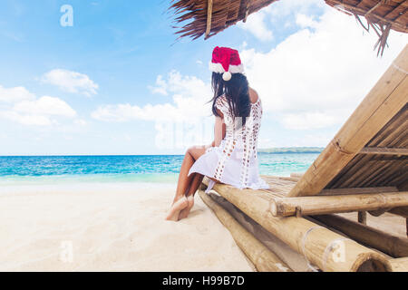 Junge Frau in Santa Claus Hut feiert Weihnachten sitzen in Chaiselongue auf tropischen Meeresstrand Stockfoto