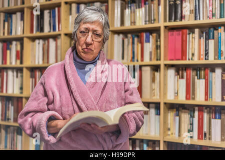 Ältere Frau liest ein Buch zu Hause. Ernsten Ausdruck. Eine verschwommene Bibliothek ist im Hintergrund. Stockfoto