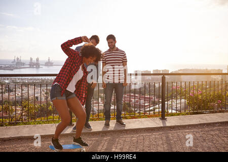 Drei coole Freunde mit einem Skateboard auf einer Brücke, alle lachen, während das Mädchen versucht, ihr Gleichgewicht auf dem skateboard Stockfoto