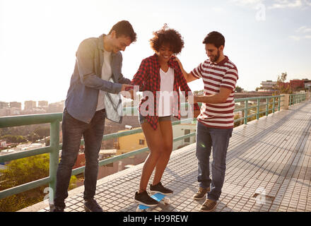 Zwei Freunde eine andere Lehre auf einer Brücke, die Männer halten das Mädchen von beiden Seiten, ihr Gleichgewicht zu halten Skateboard Stockfoto