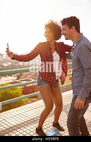 Junges Paar unter einem Selfie während balanciert, das Mädchen auf einem Skateboard und der junge hält ihre stationären Stockfoto