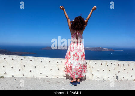 Eine Frau steht auf einer Terrasse über dem Meer mit erhobenen Händen und genießt das Gefühl der Brise, Santorini touristische Griechenland Atmosphäre Frau Arme auf Stockfoto