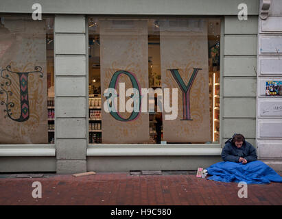 Ein Obdachloser Mann sitzt auf dem Bürgersteig von einem freudigen Shop-Schaufenster in Brighton Stockfoto