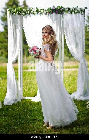Schöne blonde Braut im weißen Hochzeitskleid vor dekorierte Hochzeit Bogen, Sommerzeit, Brautstrauß Stockfoto