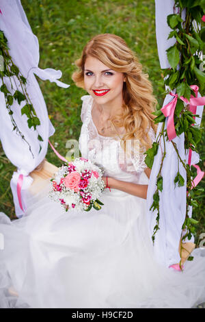 Schöne blonde Braut im weißen Hochzeitskleid schwingt sich auf dekorierten Swing und lächelt in die Kamera Stockfoto