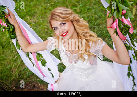 Schöne blonde Braut im weißen Hochzeitskleid schwingt sich auf dekorierten Swing und lächelt in die Kamera Stockfoto