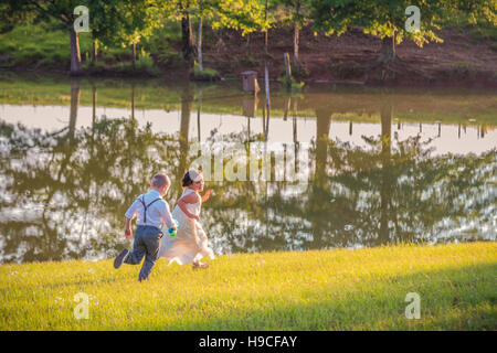 Junge Junge und Mädchen Geschwister an einem See mit gelben späten Nachmittagssonne im Feld laufen Stockfoto
