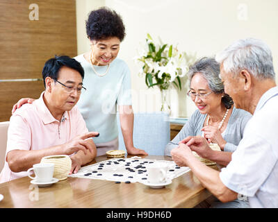 zwei ältere Männer spielen Weiqi (oder Go-Spiel) zu Hause mit ihren Frauen beobachten Stockfoto