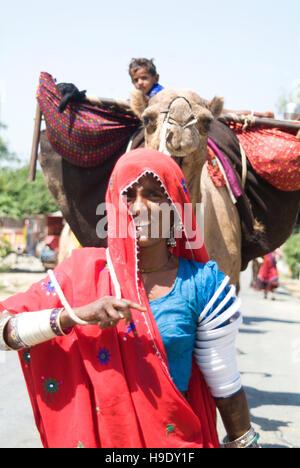 Eine nomadische Frau führt ihr Kamel vollgepackt mit weltlichen Besitztümer, darunter Kind, entlang einer Straße im südlichen Rajasthan, Indien. Stockfoto