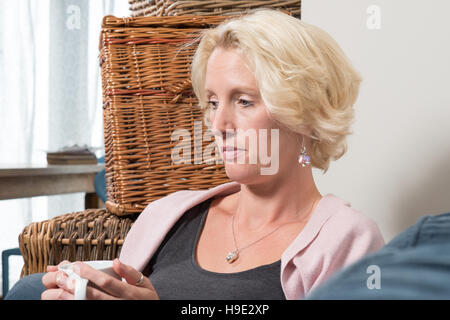 Eine blonde Frau mittleren Alters sitzt neben Korbwaren Boxen auf ein Sofa oder eine Couch, einen Becher mit beiden Händen halten und ernsthafte suchen, während sie denkt Stockfoto