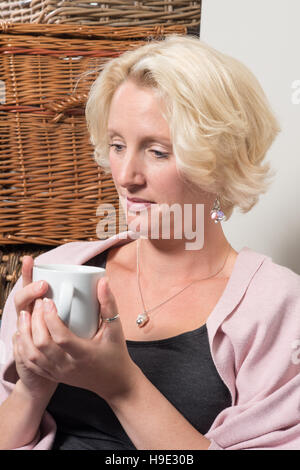 Eine blonde Frau mittleren Alters sitzt neben Korbwaren Boxen auf ein Sofa oder eine Couch, einen Becher mit beiden Händen halten und auf der Suche nach zufrieden mit einem kleinen Lächeln auf Ihr Stockfoto