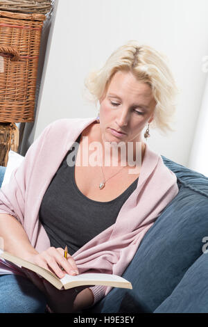 Eine leger gekleidete mittleren gealterte blonde Frau sitzt auf einem Sofa oder eine Couch Betrieb ein Bleistift und ein Buch zu lesen. Kopieren Sie Raum. Stockfoto