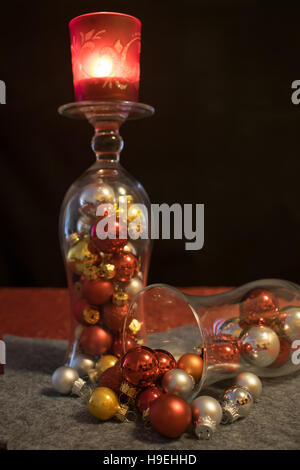 2 cocktail-Gläser gefüllt mit bunten Weihnachtskugeln auf eine graue Untersetzer. Ein Glas steht auf dem Kopf und trägt eine rote Teelichthalter mit einem brennenden Stockfoto
