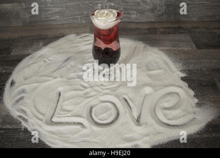 Ein Cocktailglas mit einer schwimmenden Rose und eine weiße Kerze in den Sand. Es ist das Wort Liebe in den Sand. Stockfoto