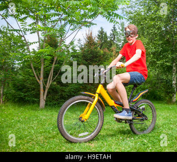 Cute teenboy tragen rote Tshirt und Schutzbrillen mit dem Fahrrad in einen Blick in die Kamera Sommerpark Stockfoto