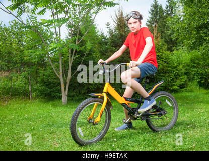 Cute teenboy tragen rote Tshirt und Brille sitzt auf einem Fahrrad in einen Blick in die Kamera Sommerpark Stockfoto