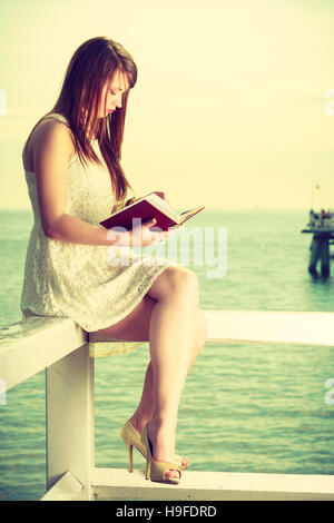 Bücher, Bücherwurm, Reisen, Freizeit Zeit Konzept. Schöne Frau im weißen Kleid sitzt auf hölzernen Hürde in der Nähe von Meer und Lesung Buch Stockfoto